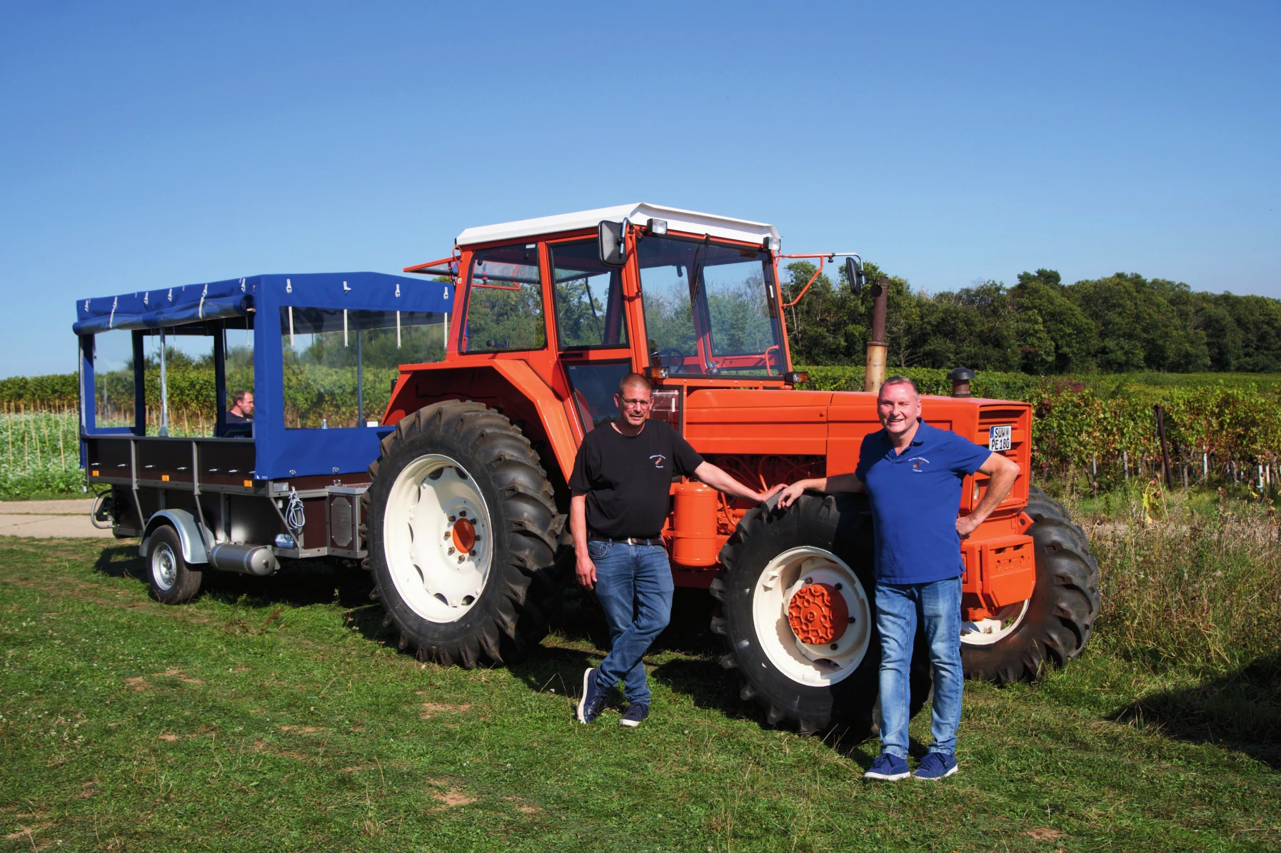 Zwei Männer vor dem roten Traktor mit blauem Planwagen in den Weinbergen